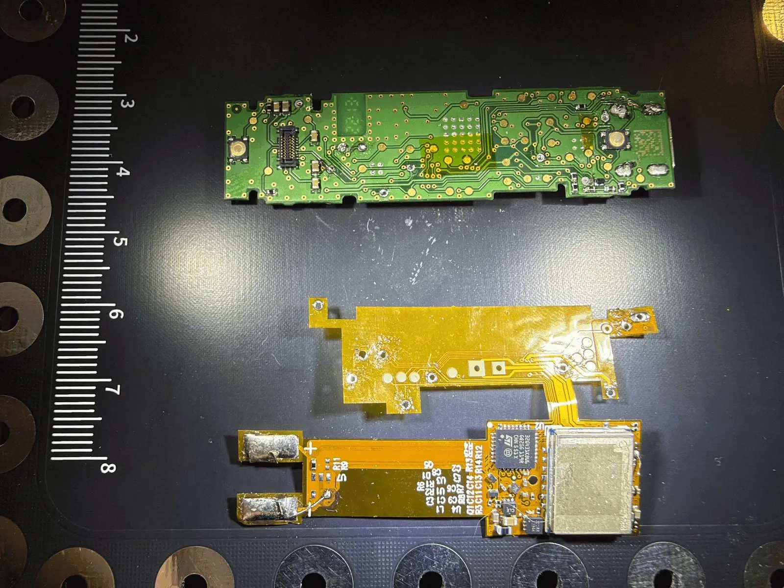 Genuine green Ledger circuit board above a separate orange flexible implant with a metal cellular module, photographed beside a ruler
